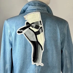 Veste en jean bleu clair avec un grand patch abstrait dans le dos. Le patch présente un motif dans les tons de noir, de gris et de crème. La veste a un col classique et une fermeture boutonnée.