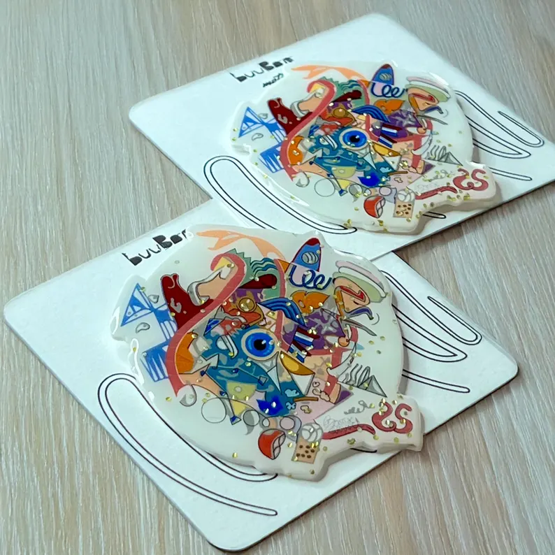 il 794xn.7018164536 bpve.jpg Broche graffiti multicolore inspirée de Paris 2024, épinglée sur une carte de présentation buuBoo aux lignes abstraites, posée sur une table en bois.
