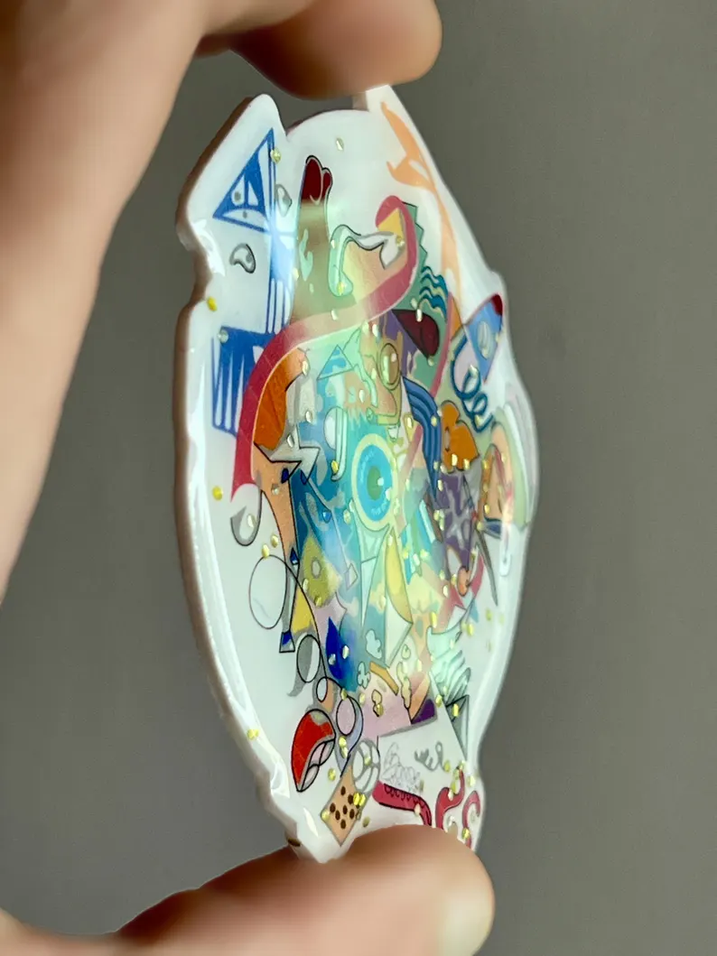 il 794xn.7066144055 paa4.jpg Gros plan d’une broche graffiti multicolore inspirée de Paris 2024, tenue entre les doigts pour montrer les détails du dessin et la finition brillante en résine époxy. buuBoo.