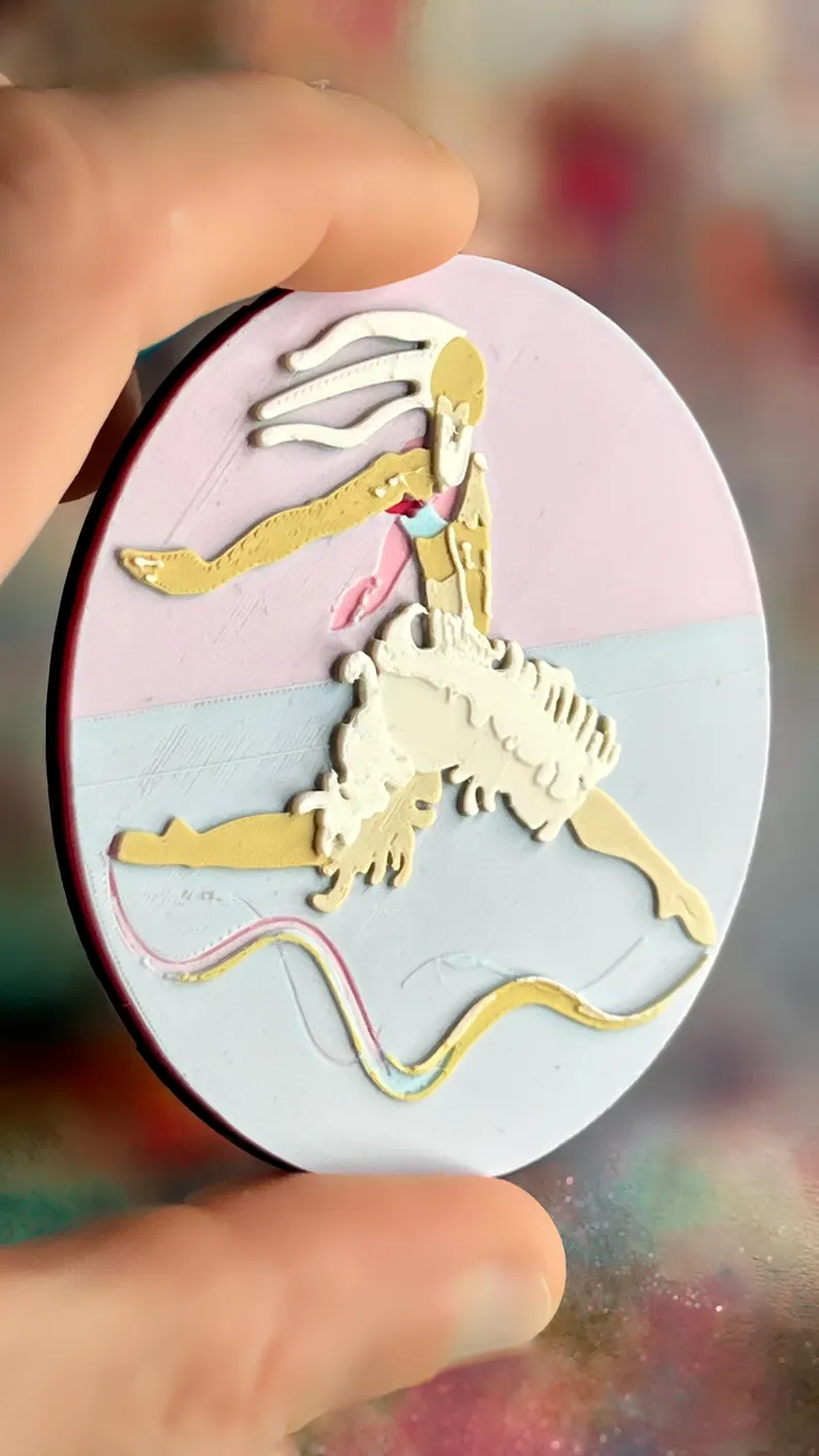 il 794xn.8344682015 lyk0.jpg Broche ronde Danseur-Cygne présentée en main, relief imprimé en 3D avec palette pastel effet nuage, personnage aux dégradés beige et blanc et fond rose bleu doux. Marque buuBoo.